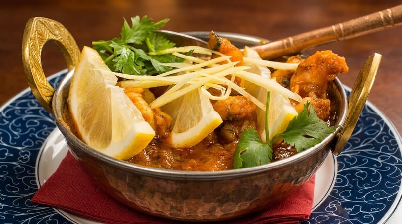 Indian curry in a copper bowl, garnished with lemon wedges, ginger strips, and cilantro.