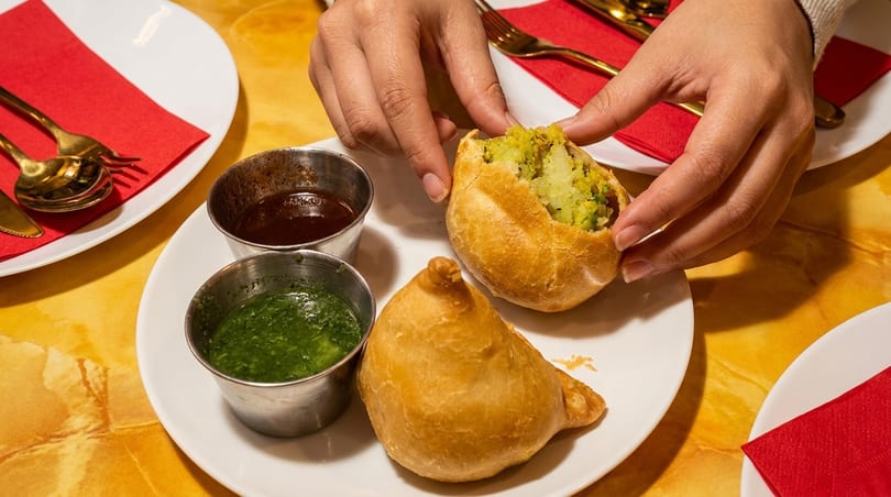 Hands opening a samosa to reveal its potato and pea filling, with chutneys on a white plate.