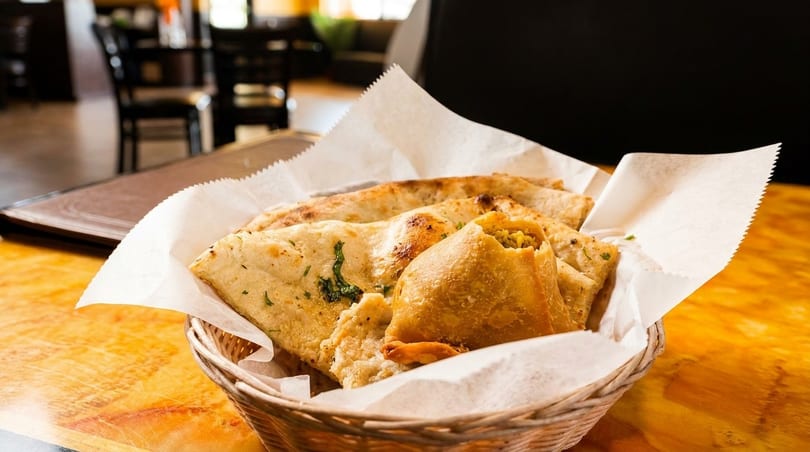 A woven basket filled with naan bread and a samosa, on a wooden table in a restaurant.